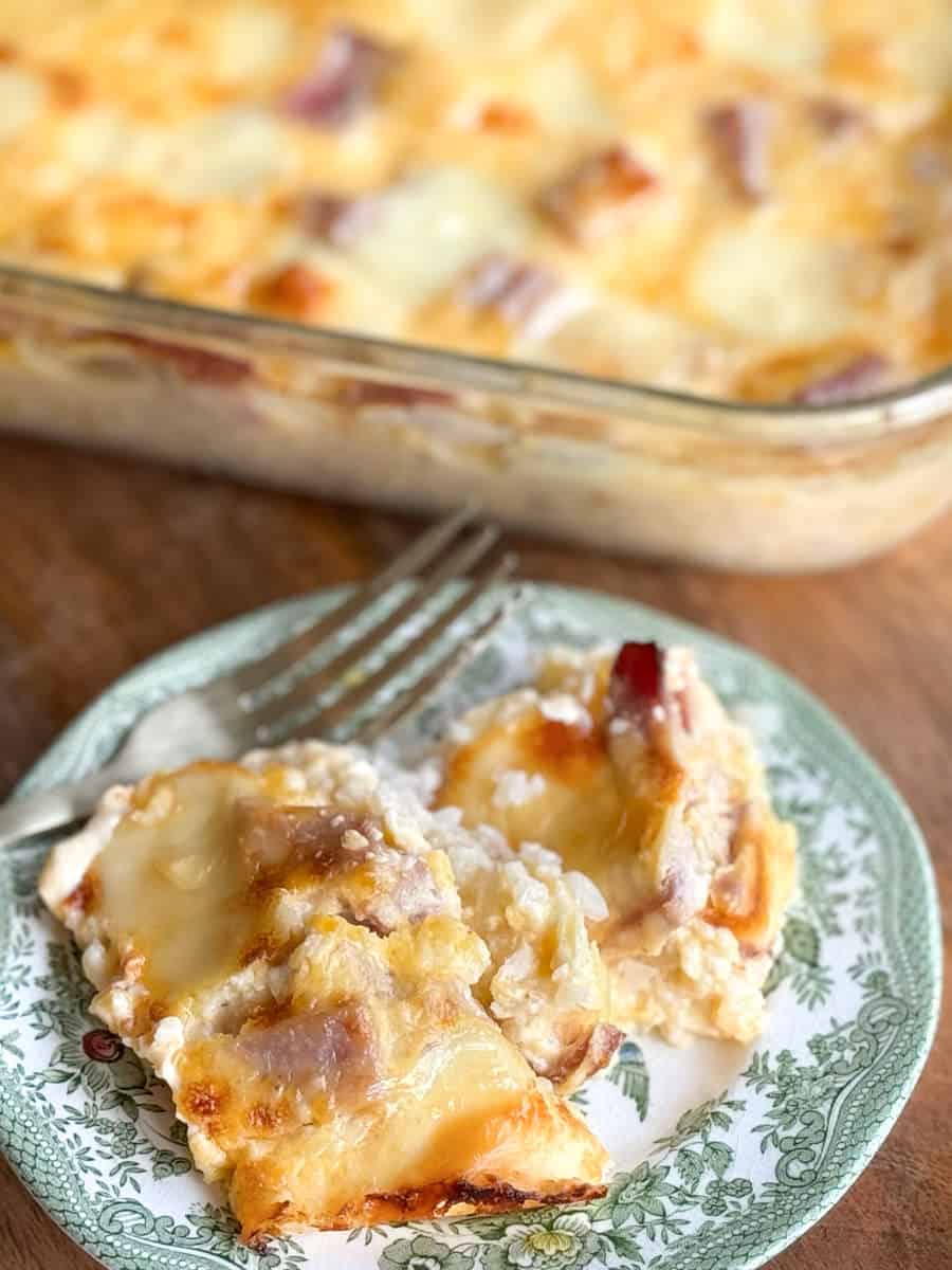 A close-up of a white and green floral-patterned plate containing a serving of a baked dish with melted cheese and pieces of ham. A glass baking dish with the same baked dish is visible in the background along with a partially visible fork resting on the plate.