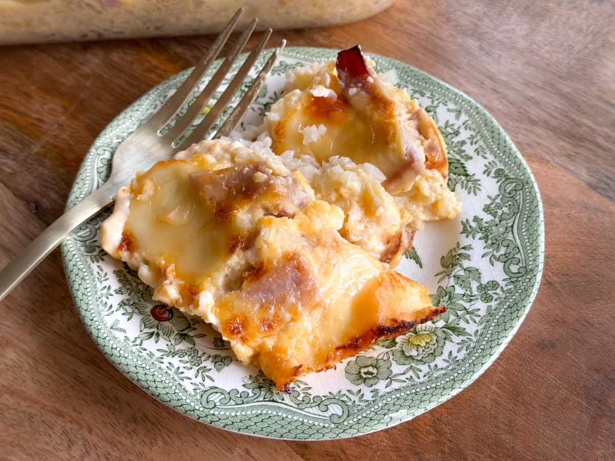A plate with two slices of a cheesy baked dish, possibly scalloped potatoes or lasagna, served on a decorative green and white plate. The dish appears creamy with a golden-brown top. A fork is placed on the plate next to the slices.