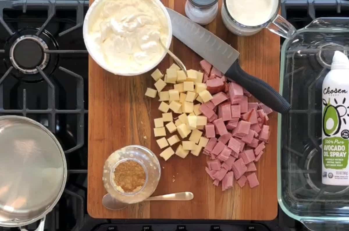 A wooden cutting board on a stove holds a knife, cubed ham, and cheese. Surrounding it are a glass jar with a brown substance, two open jars with white contents, a can of avocado oil spray, a stainless steel pot, and a clear glass baking dish.