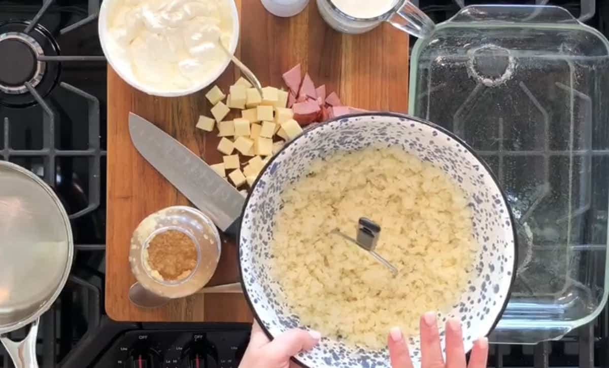 A person is preparing food on a wooden board placed on a stovetop. On the board are a large bowl of mashed potatoes, diced cheese and ham, a knife, a bowl of sour cream, a cup of milk, and a clear baking dish. The person is holding the bowl of mashed potatoes.