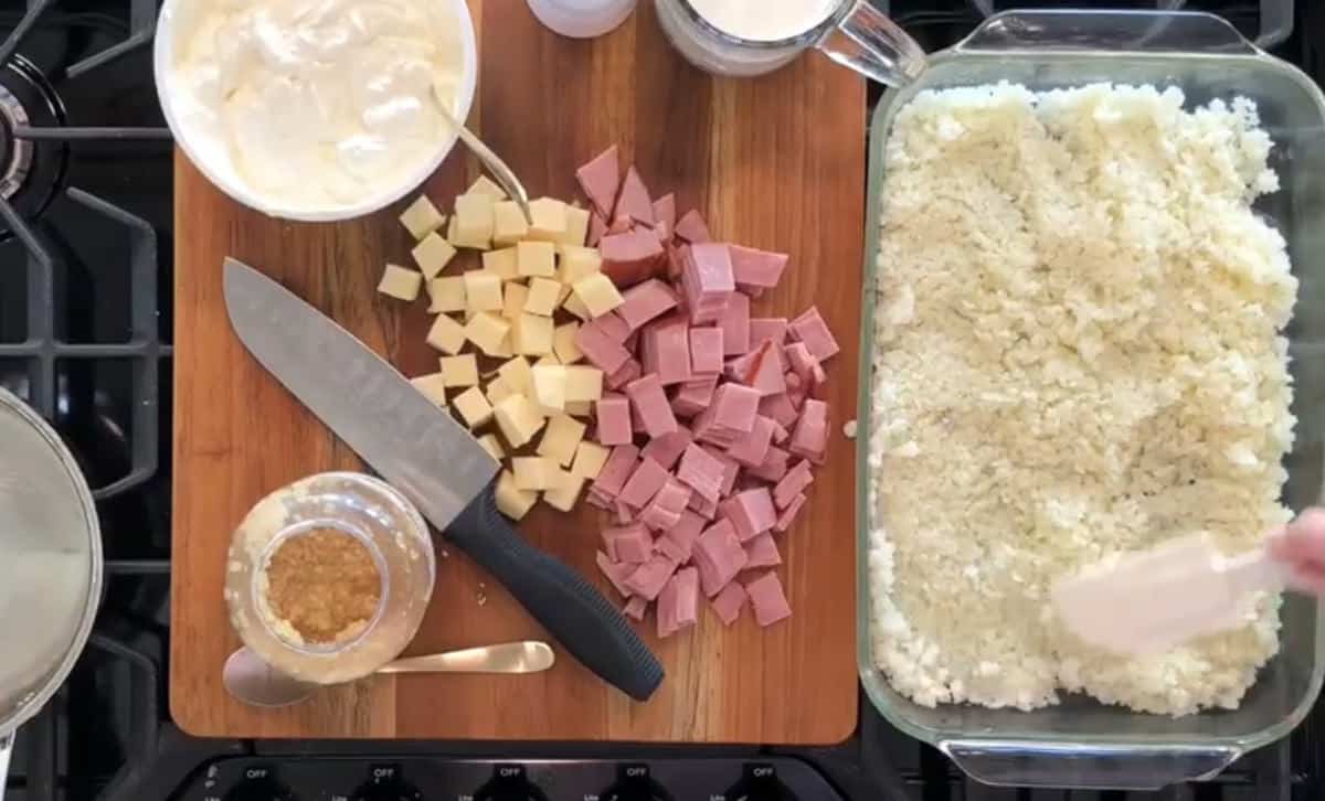 A cutting board with cubed ham and cheese, a large knife, a bowl of cream, a glass measuring cup, and a jar of minced garlic. A hand is spreading a mixture in a glass baking dish on the right side of the image.