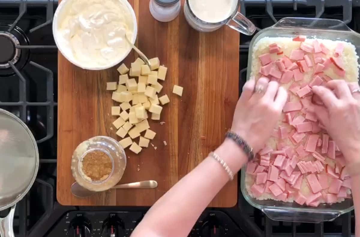 A person is seen preparing a dish on a wooden cutting board. There are cubes of cheese, a bowl of creamy mixture, a glass jar with a brown substance, and chopped ham in a casserole dish. The person's hands are arranging ham pieces in the dish.
