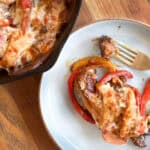 A skillet dish of baked bell peppers filled with melted cheese and meat served on a wooden table beside a plate with a partially eaten pepper and a fork.
