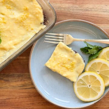 A slice of lemon bar with a crumbly base and lemon curd topping on a blue plate beside fresh lemon slices, a mint leaf, and a fork. a larger tray of lemon bars in the background rests on a wooden table.