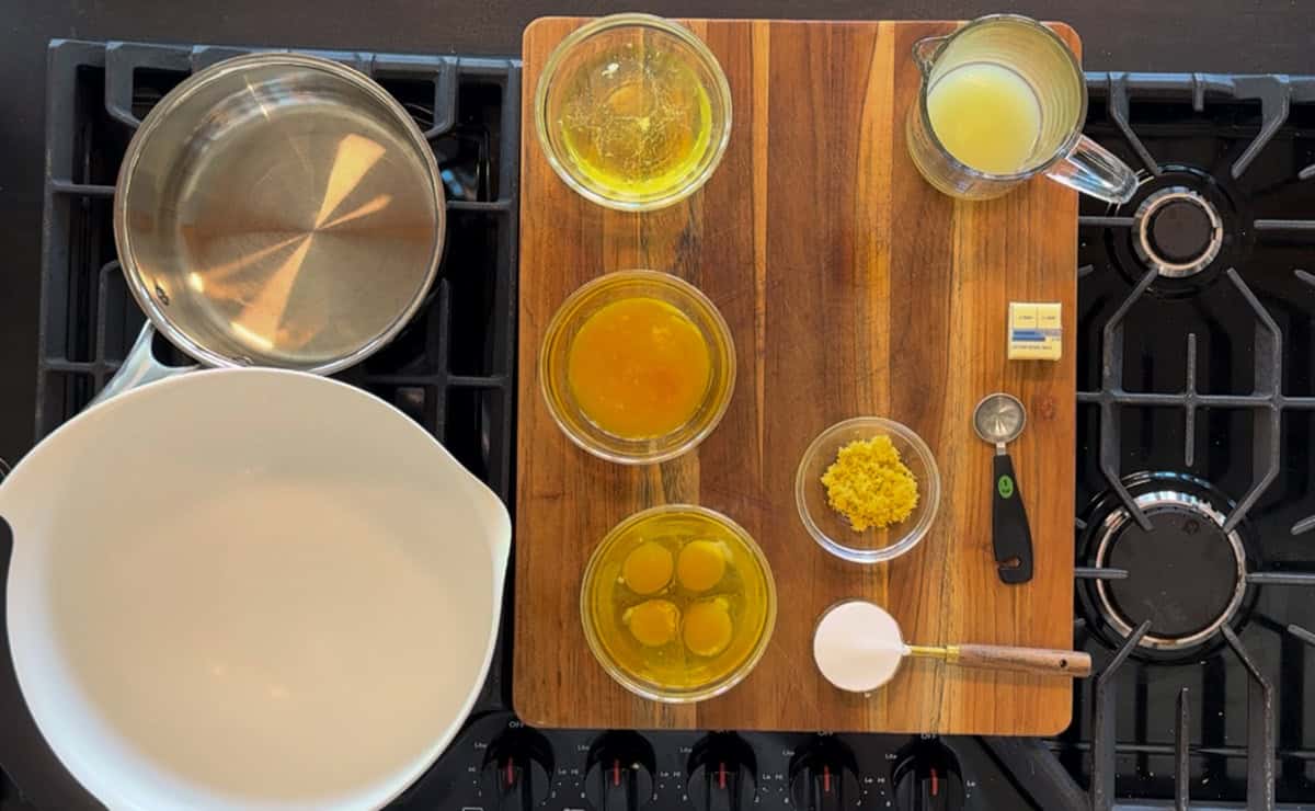 Top view of a kitchen countertop setup for cooking, featuring a wooden cutting board surrounded by ingredients and utensils, including a bowl, pots, eggs, and lemon zest. a stove burner is visible.