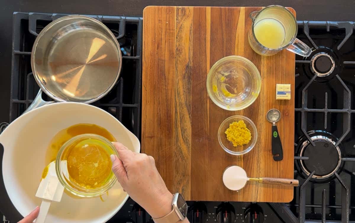 A person is pouring egg whites into a bowl on a stovetop, with ingredients like flour, sugar, and lemon zest neatly arranged on a wooden cutting board nearby.