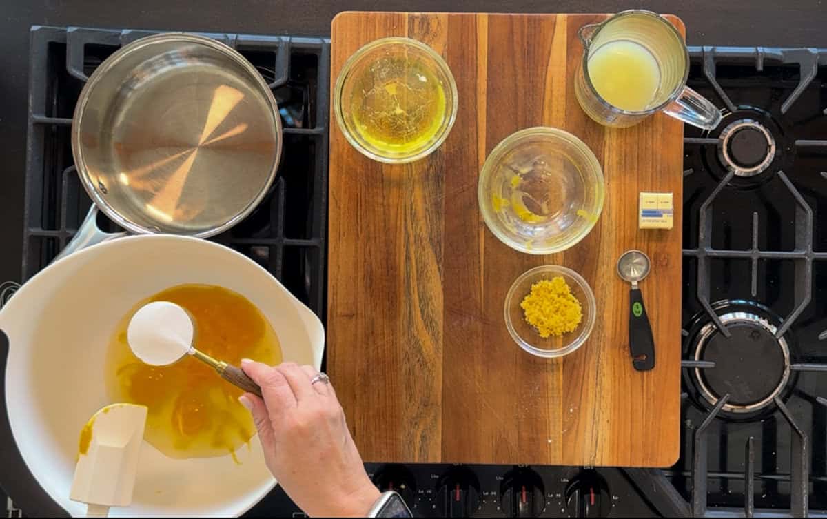 Overhead view of a kitchen stove workspace with a person pouring liquid into a bowl, surrounded by ingredients such as oil, lemon zest, and utensils on a wooden board.
