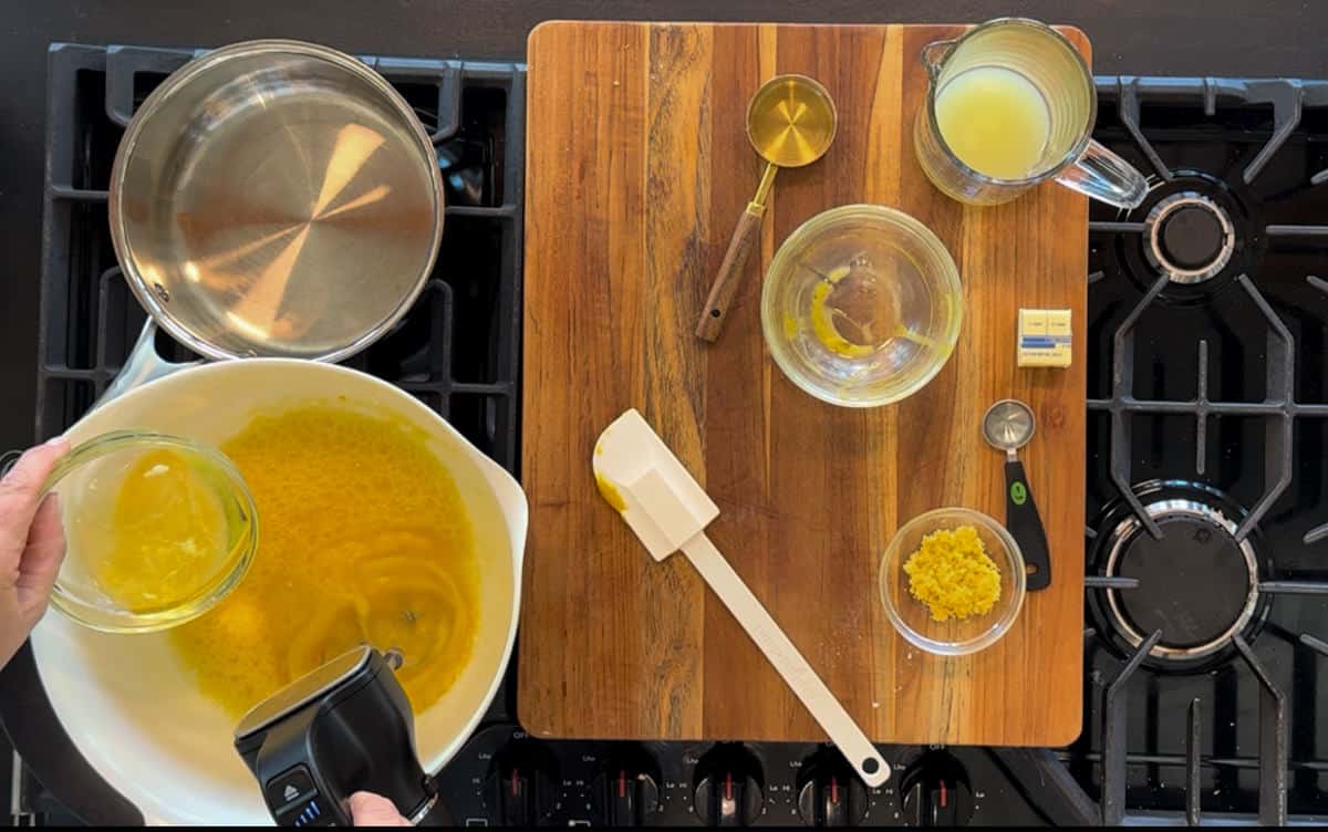 Overhead view of a cooking scene on a stove featuring a wooden cutting board, bowls with lemon zest and juice, a silicone brush, a melted butter container, and an open butter pack.
