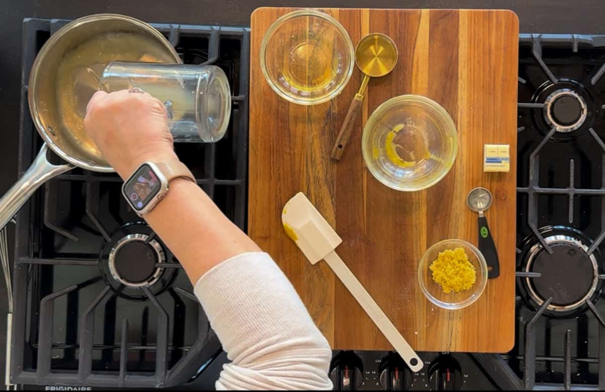 A person pours liquid from a saucepan into a glass bowl on a stove, surrounded by ingredients and cooking utensils on a wooden cutting board.