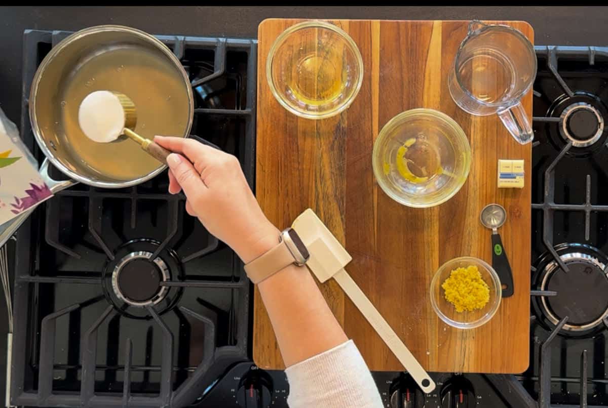 A person's hand stirs a pan on a stove, surrounded by neatly arranged ingredients including lemon zest and oil, on a wooden cutting board in a well-organized kitchen.