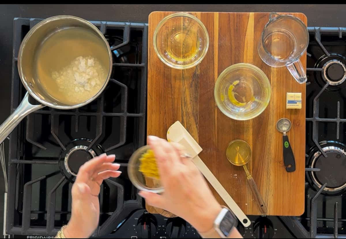 Overhead view of a cooking setup on a stove, featuring a pot with liquid, wooden cutting board with ingredients arranged in bowls, and hands holding lemon zest.