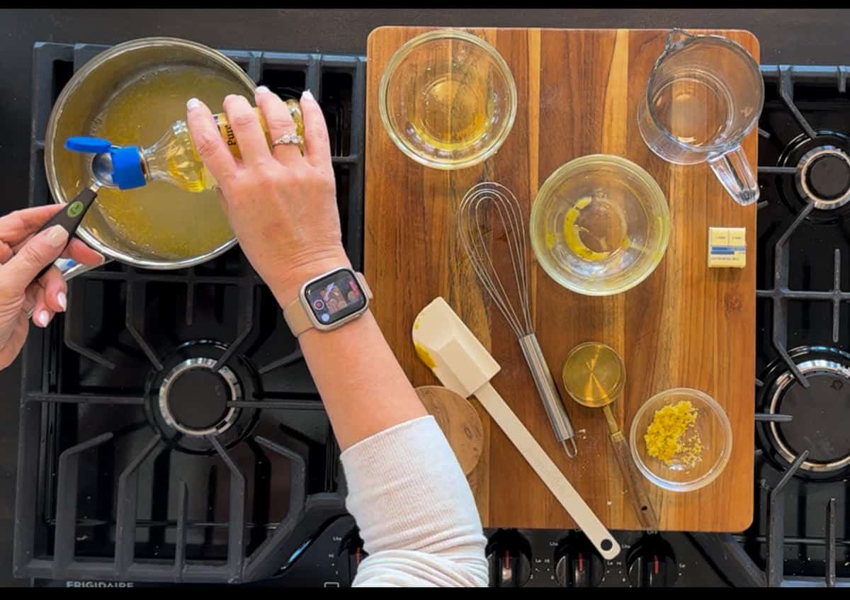 Overhead view of a person's hands pouring oil from a bottle into a pan on a stove, surrounded by various cooking utensils and ingredients arranged neatly on a wooden counter.