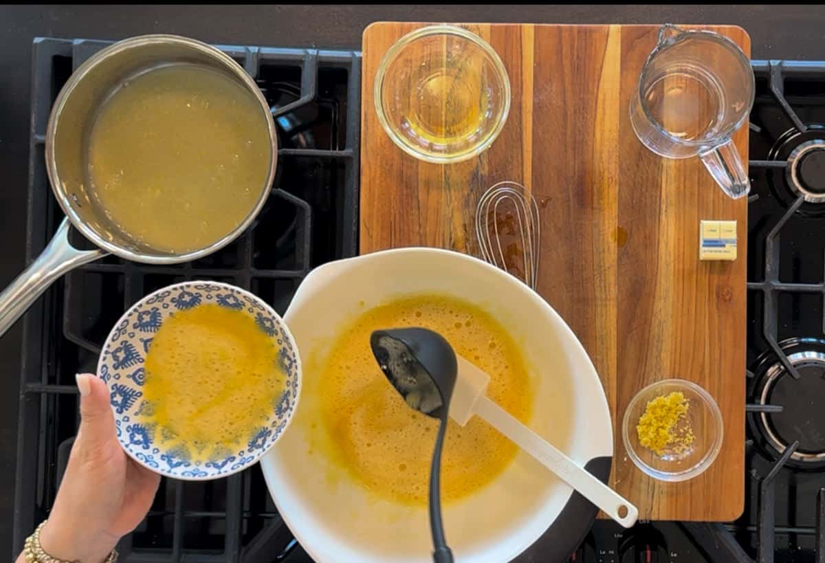 An overhead view of a cooking scene on a stove featuring a hand holding a bowl with whisked eggs, a saucepan with soup, two empty glasses, and various ingredients laid out on a wooden board.