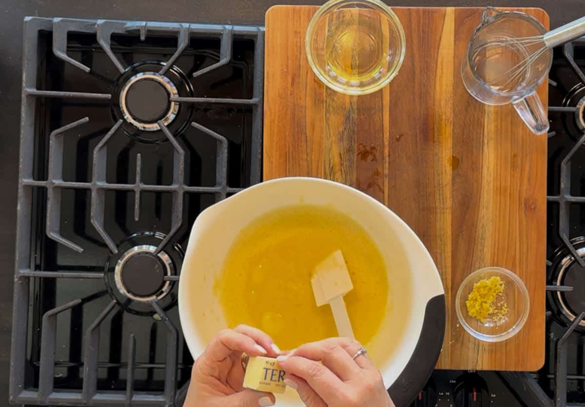 Overhead view of a kitchen counter with a person's hands stirring a bowl of melted butter, next to a stove, a glass jug, two glasses with yellow liquid, and a small bowl of grated zest.
