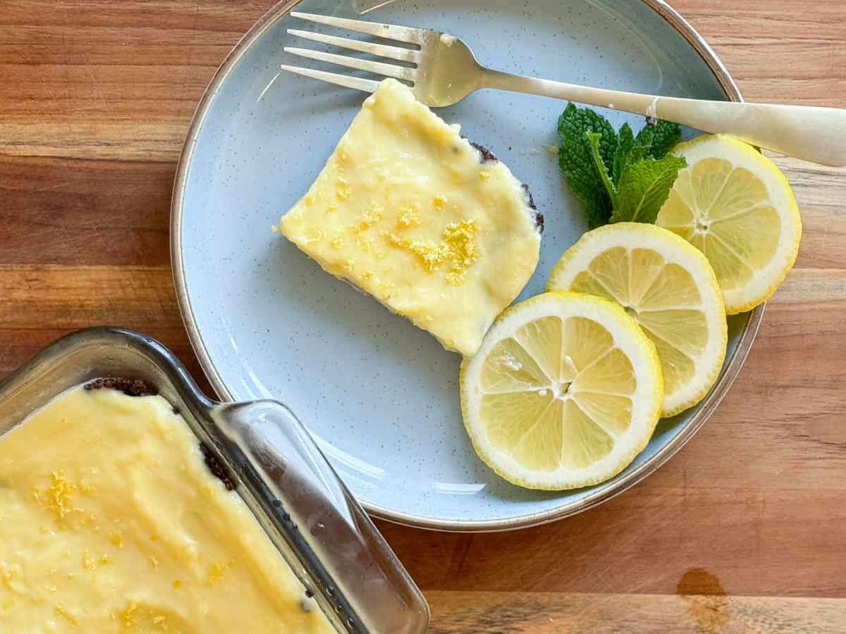 A slice of lemon cake with a lemon glaze, accompanied by sliced lemons and fresh mint leaves, presented on a blue plate with a wooden fork. a baking dish with more cake is partly visible.