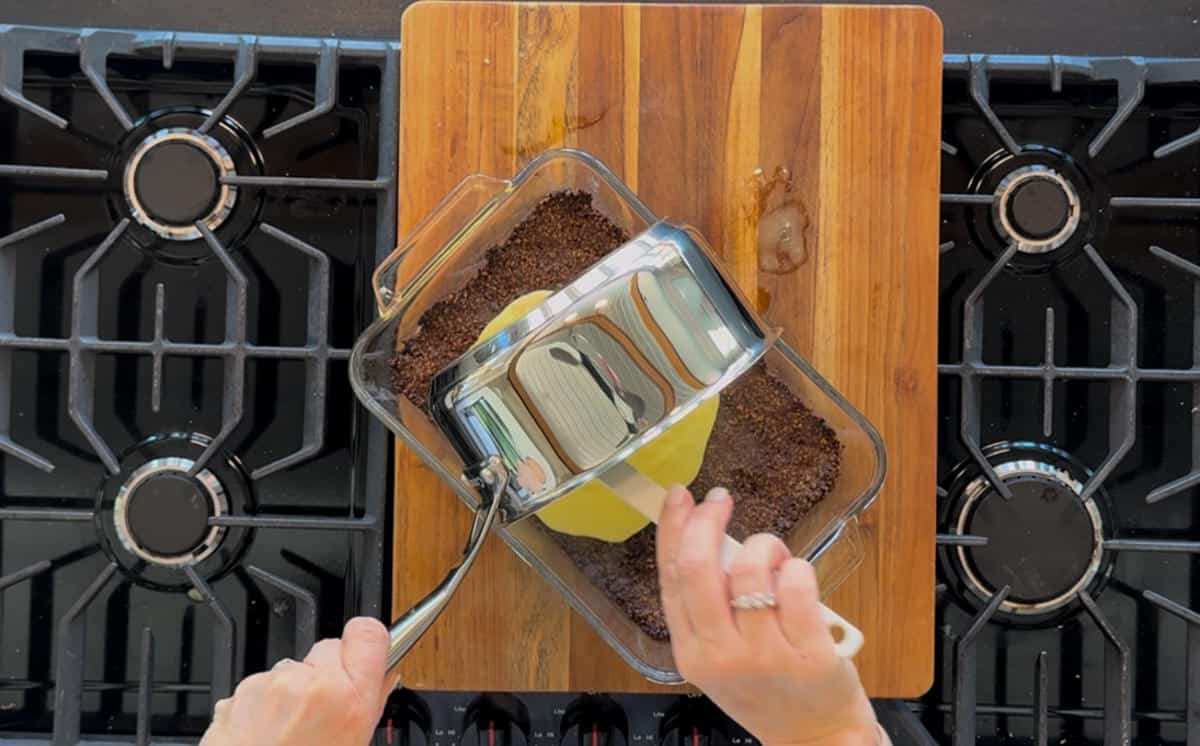 A person uses a metal spoon to blend the lemon bar mixture over the crust in a clear glass mixing bowl on a wooden cutting board over a stove top.