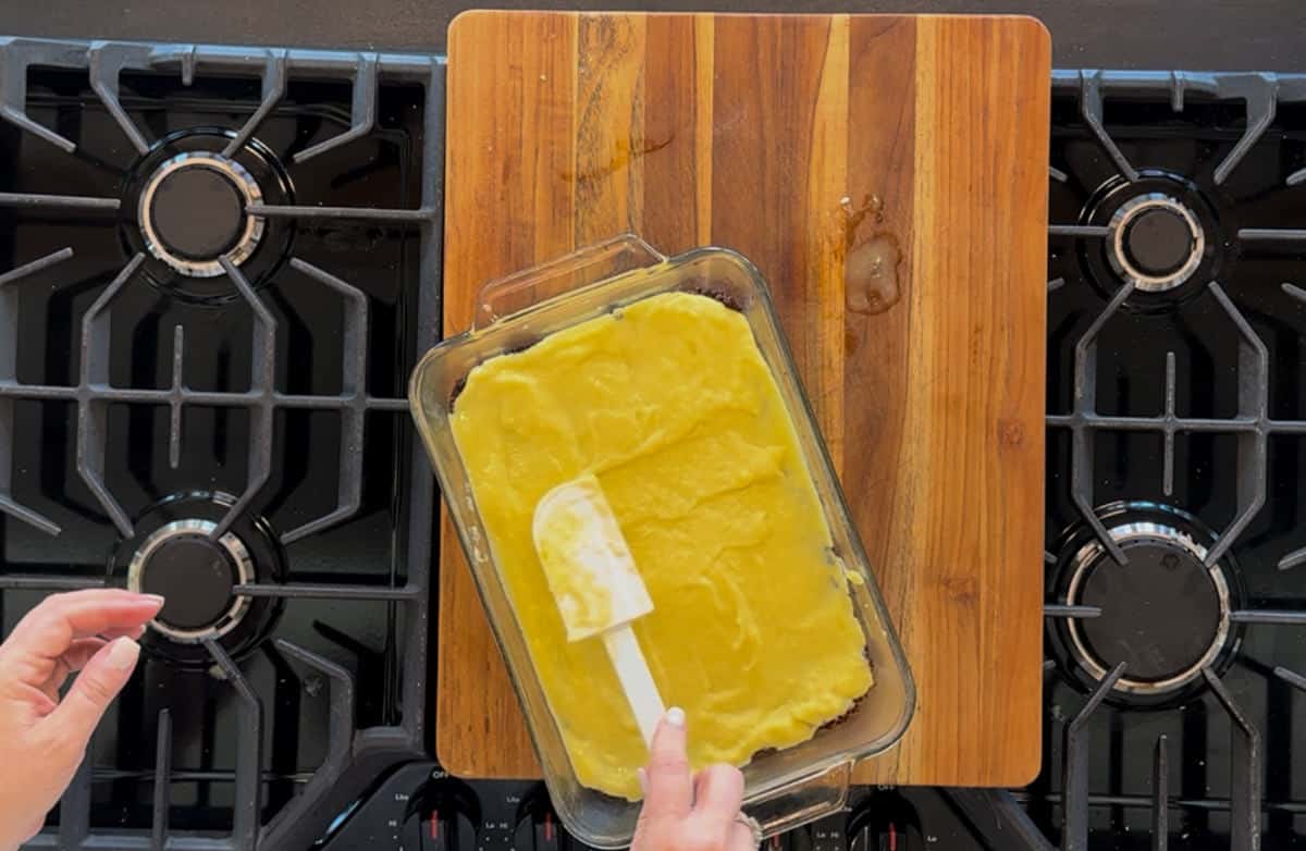 A person spreads a yellow mixture in a glass baking dish on a wooden cutting board, placed on a stove top. there are traces of the mixture on the board.