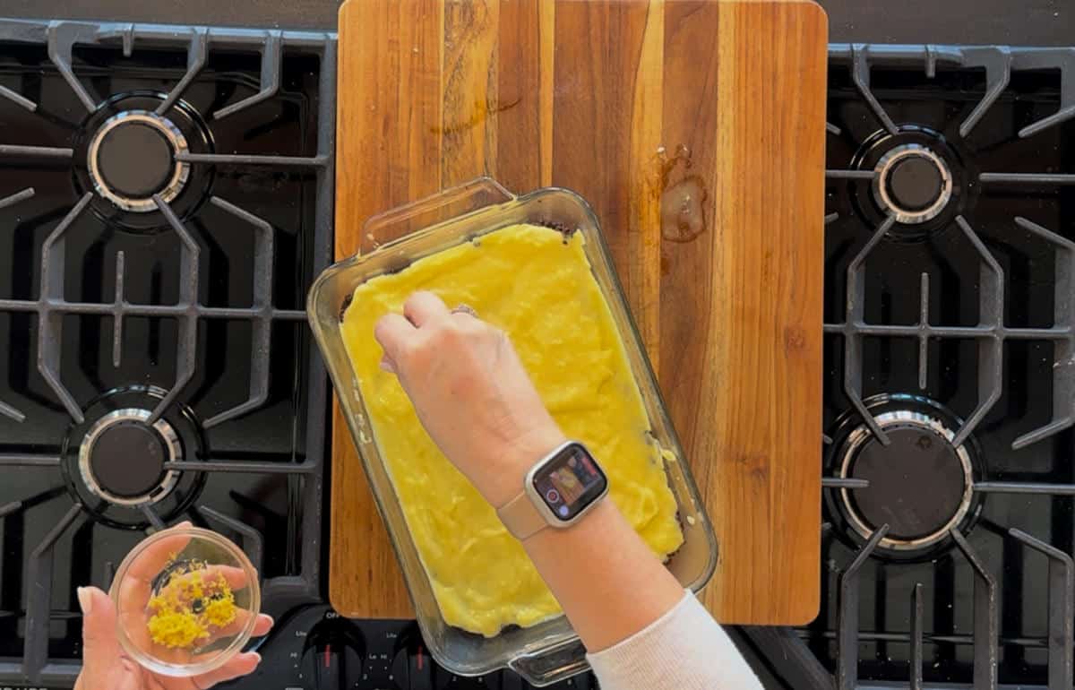 A person wearing a smartwatch is sprinkling yellow lemon zest over a baking dish containing a yellow mixture, resting on a wooden cutting board beside a stovetop.