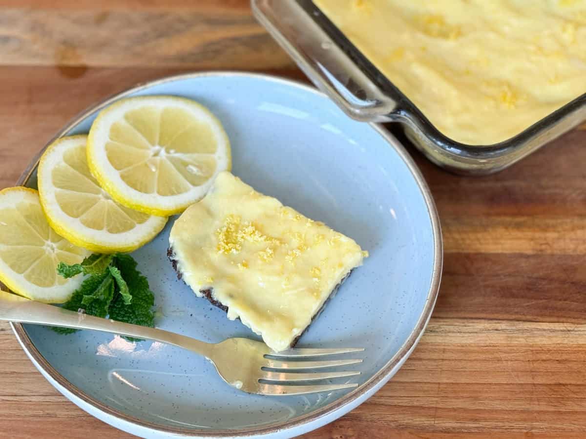 A slice of lemon dessert on a blue plate, garnished with lemon slices and mint, served with a fork. a baking dish with more dessert visible in the background, all on a wooden table.