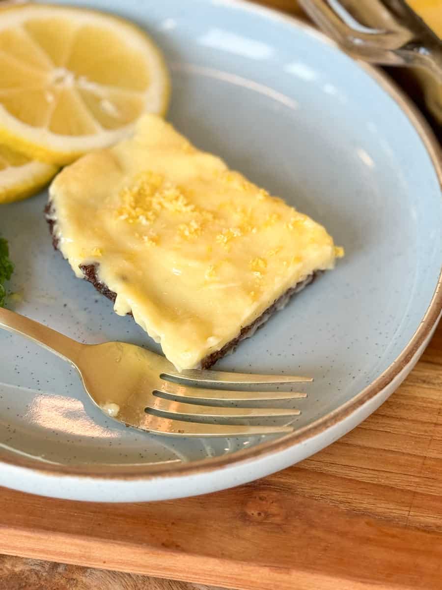 A slice of lemon dessert with a creamy yellow topping and crumbly base on a blue plate, garnished with lemon slices. a fork rests beside the plate.