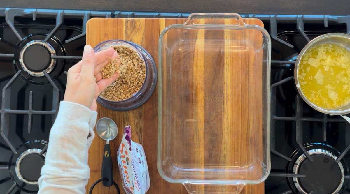 A person's hand sprinkling a jar of crushed pecans into a glass baking dish on a stove, next to a wooden cutting board and a pot of melted butter.