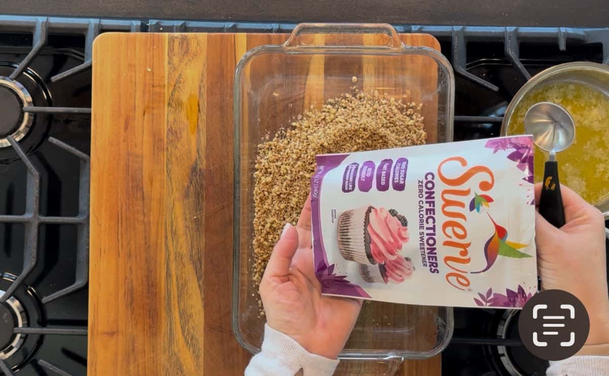 A person's hands are pouring crushed pecans from a saxon confectioners bag into a glass baking dish next to a wooden cutting board, all set on a stovetop.