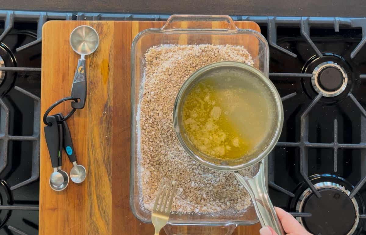 Hands pouring melted butter from a small saucepan into a glass baking dish filled with crumbs, on a stove. measuring spoons rest beside the dish.