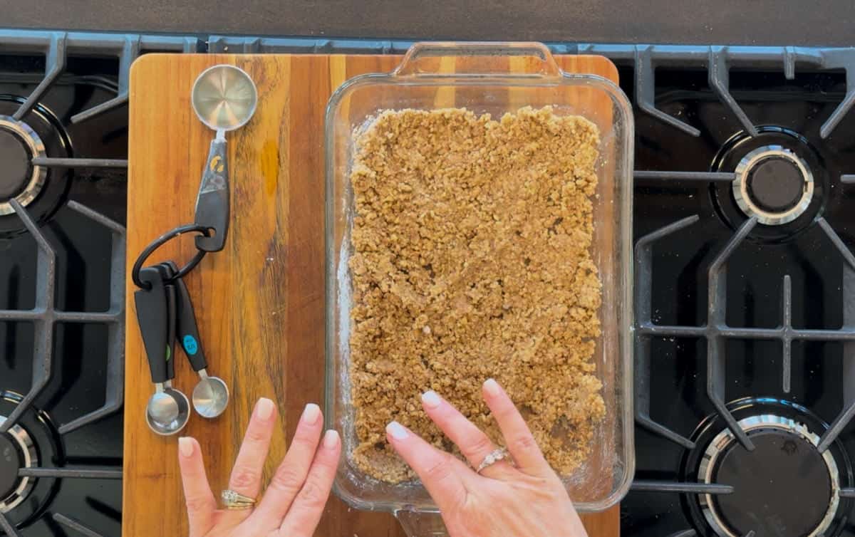 Top-down view of hands pressing a crumbly brown mixture into a glass baking dish on a wooden cutting board next to kitchen utensils, set on a stove.