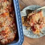 A baked casserole dish filled with a cheesy tomato mixture sits next to a plate with a serving of the casserole. The plate also has a fork. The casserole is topped with breadcrumbs and shows visible layers of tomato slices and cheese.