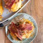 A plate holds a serving of cooked chicken wrapped in bacon, resting next to a fork. The dish appears cheesy and seasoned, with an additional portion visible on a serving plate in the background. The food is placed on a wooden surface.