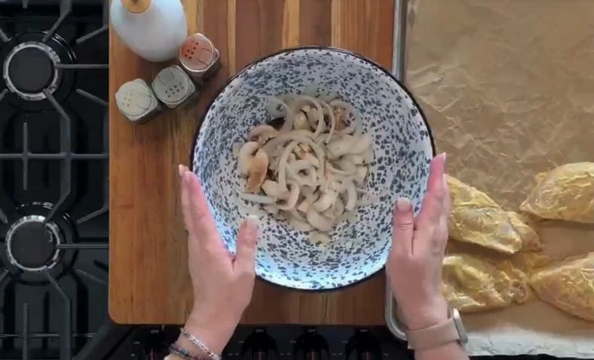 A person is holding a bowl of sliced mushrooms over a wooden cutting board. Nearby are three spice containers, a white jar, and a baking tray with uncooked seasoned chicken pieces on parchment paper. The background shows a stovetop.