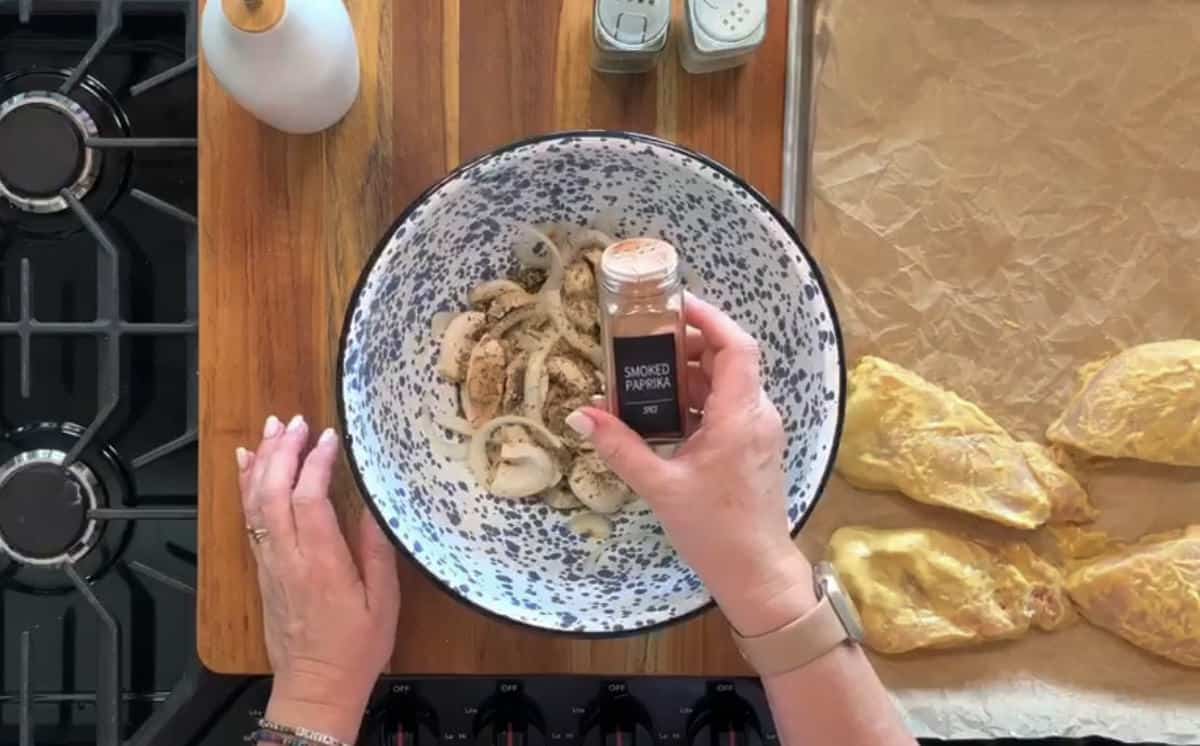 A person is preparing ingredients on a wooden countertop. They are holding a container of smoked paprika over a bowl of sliced mushrooms and onions. A baking tray with raw seasoned chicken breasts and various spices are also visible. Part of a stove is on the side.