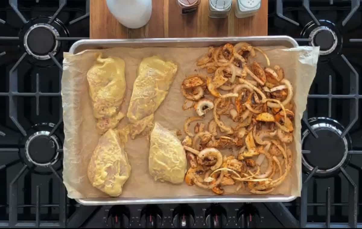 A baking sheet placed on a stove top holds four seasoned chicken breasts on one side and a mixture of sliced onions and spices on the other side. The sheet is lined with parchment paper. In the background, a wooden cutting board with condiments is visible.