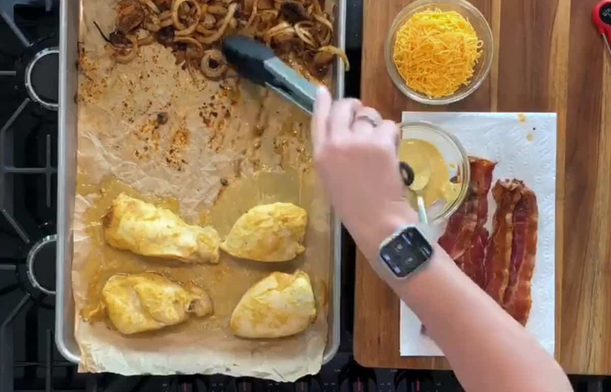 A hand using tongs handles cooked onions on a baking sheet. The sheet also has cooked chicken pieces. A wooden board beside it holds grated cheese, cooked bacon strips on a paper towel, and a bowl of sauce. A person’s wrist wearing a smart watch is visible.