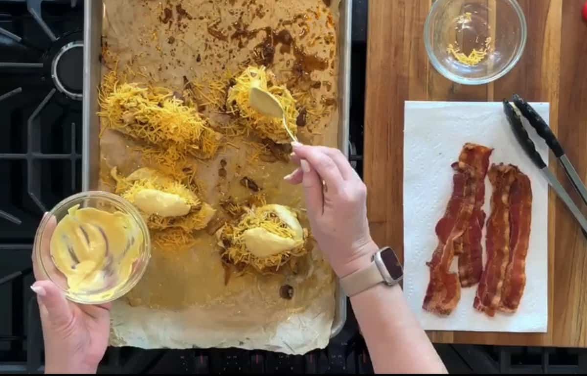 A person is using a spoon to spread a yellow sauce over shredded cheese-covered stuffed chicken breasts on a parchment-lined baking sheet. Three cooked bacon strips are placed on a paper towel, and empty bowls and tongs are on a wooden cutting board.