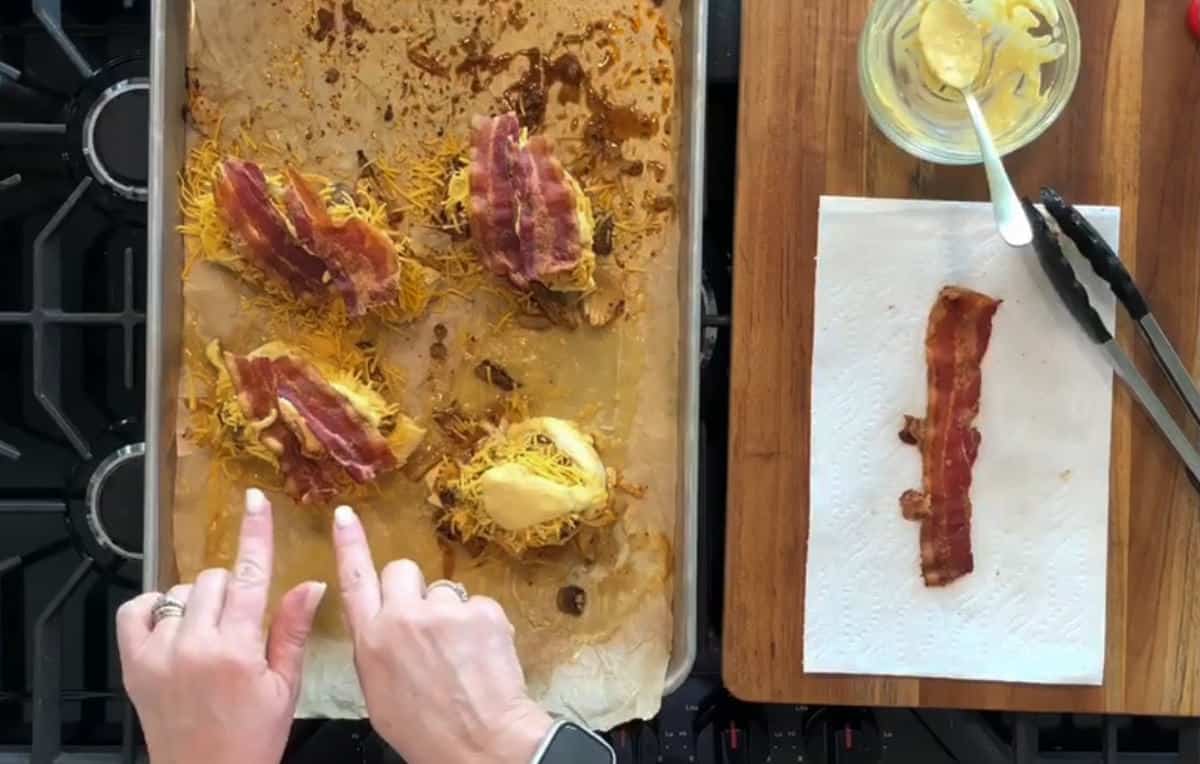 A person arranging bacon slices on top of shredded cheese and other toppings on baked potato skins on a parchment-lined baking tray. Nearby, there is a wooden cutting board with a bowl of butter, a pair of tongs, and a paper towel with a bacon strip.