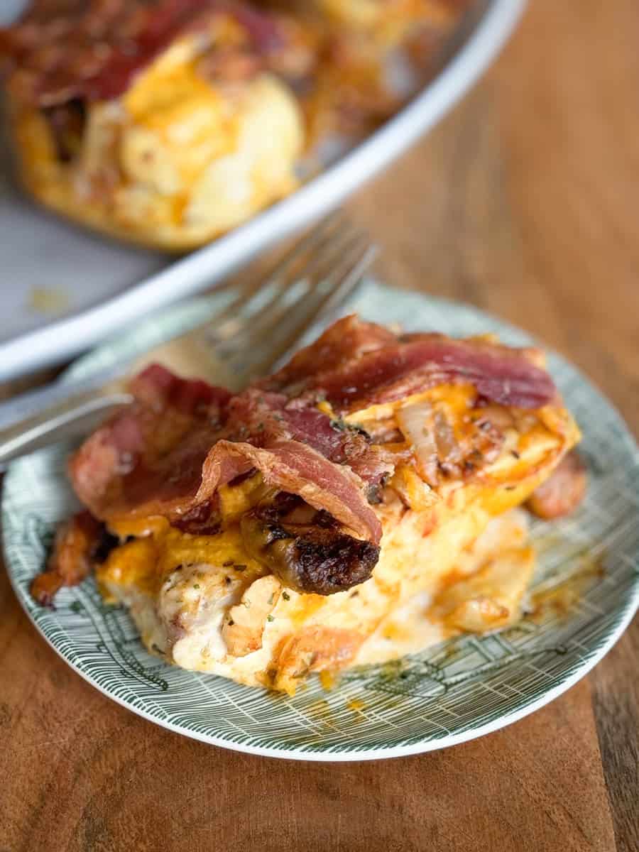 A plate with a serving of chicken topped with melted cheese, crispy bacon strips, and caramelized onions. A fork rests on the edge of the plate. The background shows part of another dish with more food, all set on a wooden table.