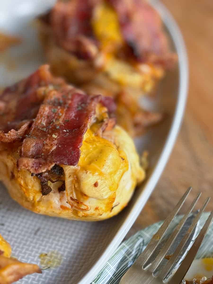 Close-up of a plate with bacon-wrapped stuffed chicken breasts. The dish is placed beside a fork on a wooden surface. The chicken is topped with melted cheese and seasoned with herbs.