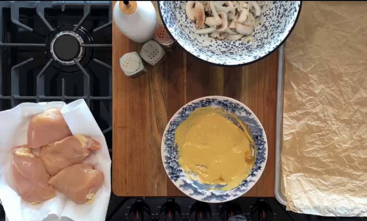 A kitchen counter with raw chicken thighs on a paper towel, a bowl of sliced mushrooms, and a bowl of mustard sauce. To the right, there is a parchment-lined baking sheet. The scene is set on a wooden surface next to a stovetop.
