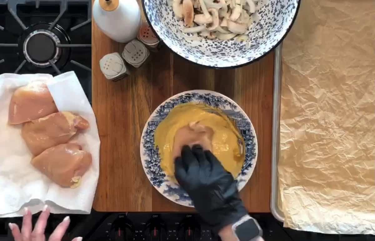 Person wearing a glove dips a raw chicken breast into a bowl of yellow batter. Nearby are more chicken breasts on a paper towel, a bowl with sliced mushrooms, and a baking tray lined with parchment paper. Various seasonings are nearby on the countertop.