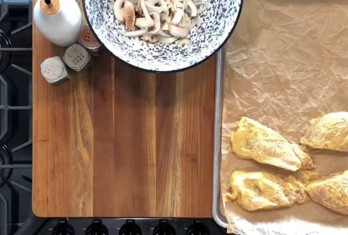 Overhead view of a wooden countertop with a gas stove. On the left, a bowl of sliced mushrooms and a bottle of oil are next to salt and pepper shakers. On the right, a baking tray with three seasoned raw chicken breasts on parchment paper.