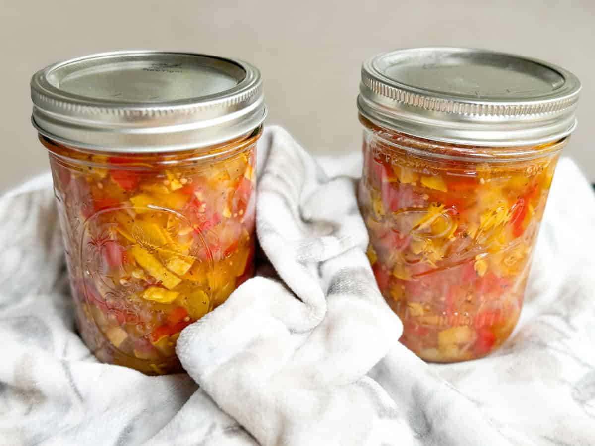 Two glass jars filled with homemade vegetable relish are placed on a white, patterned cloth. Each jar has a silver metal lid, and the relish inside consists of various chopped vegetables in a liquid mixture.