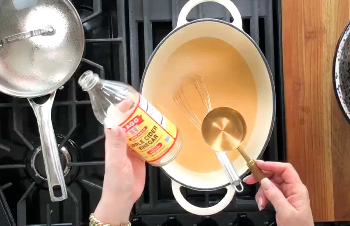 A person is holding a bottle of apple cider vinegar in their left hand and a measuring cup in their right hand, ready to pour the vinegar into a white pot on a stovetop. A metal pan with a lid is on another burner to the left.