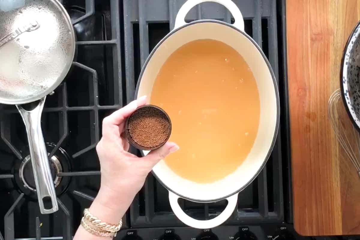 A hand is holding a small container of spices over a white pot filled with a yellowish liquid, placed on a black stovetop. There is a stainless steel pot with a lid on the stove beside the white pot. A bowl and a whisk are on the wooden countertop.