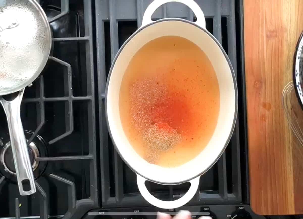 A white Dutch oven sits on a stovetop burner, containing a simmering liquid with reddish and brownish hues. There is a small saucepan with a lid on an adjacent burner. Part of a wooden countertop and a metal whisk are visible on the right side.