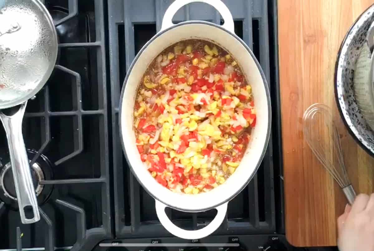 A white pot on a stove contains a mixture of chopped vegetables, including yellow and red bell peppers and onions. To the right, a whisk and a mixing bowl are partially visible on a wooden countertop. Another pot with a lid is on the left burner.