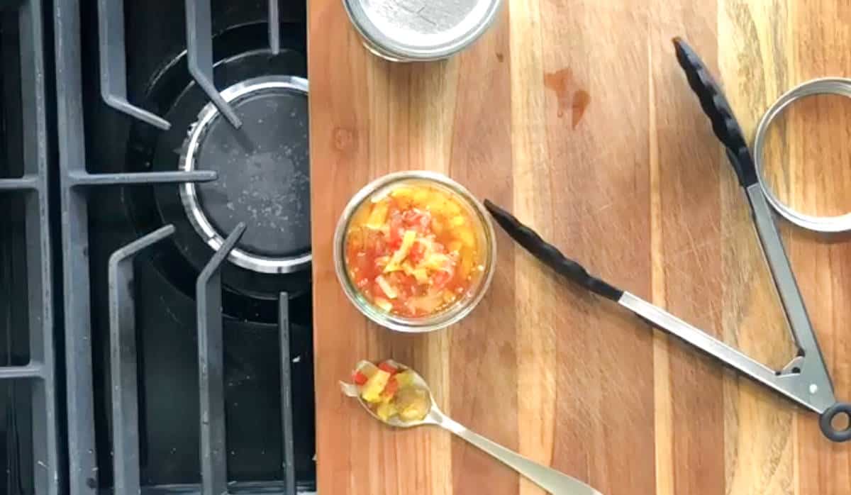 A jar of diced tomatoes and peppers is placed on a wooden countertop next to a spoon and a pair of kitchen tongs. A stove burner is visible on the left side of the image.