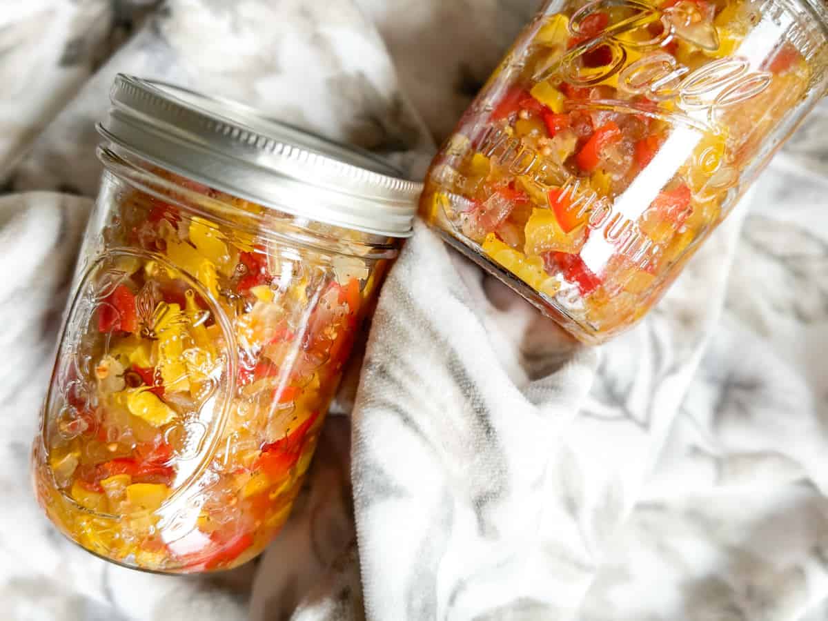 Two mason jars filled with a colorful mix of chopped pickled vegetables, featuring red and yellow peppers. The jars are placed on a soft, patterned fabric background. Both jars are sealed with metal lids.