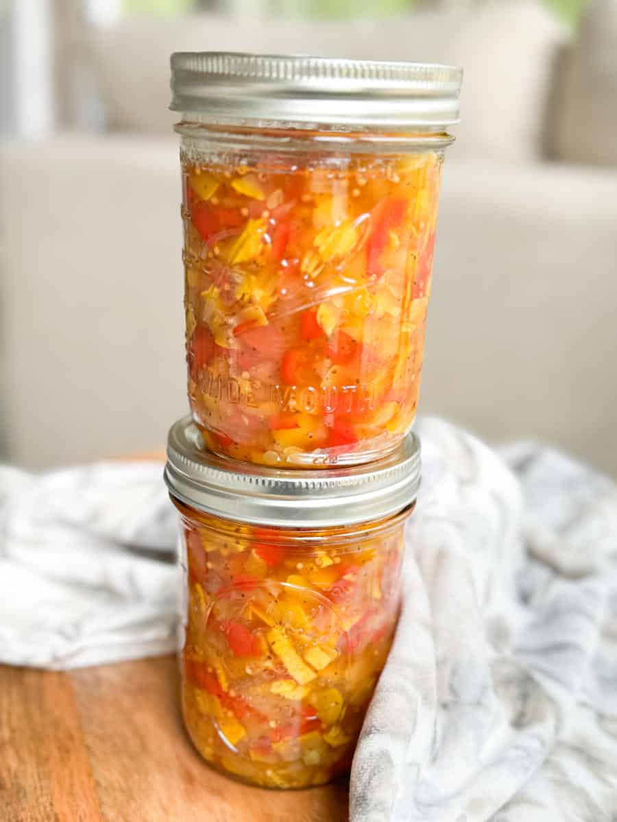 Two sealed mason jars filled with a colorful mixture of pickled peppers sit stacked on a wooden surface. The jars contain a blend of red, yellow, and green peppers in brine. A light-colored cloth is partially visible in the background.