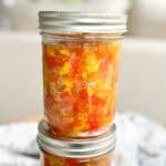 A close-up image of two glass mason jars filled with a colorful mixture of chopped vegetables, likely a homemade relish or salsa. The jars are sealed with metal lids and the top jar is slightly angled, resting on a blurry background.